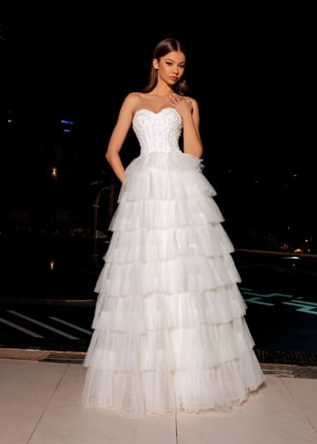 In the dimly lit night, a woman stands elegantly, her figure highlighted by the strapless white ALICE NC1045 gown. The dress features a fitted lace bodice and a tiered tulle skirt that gracefully reaches the floor. With long, straight hair and a confident, poised expression, she embodies the grace of ALICE NC1045 perfectly. Angels Formal Wear