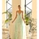 A woman stands in an elegant, sunlit room wearing a long, flowing, light blue ALEXIS NC1070 dress adorned with floral patterns. The room features tall windows with a crisscross design, large potted plants, and ornate railings, creating an opulent backdrop. Angels Formal Wear