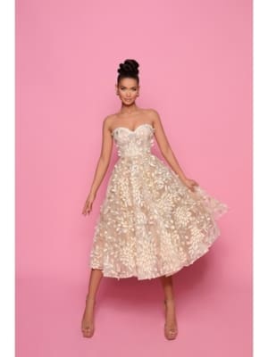 A person stands against a pink background wearing a strapless beige RILEY NP145 dress adorned with floral patterns. They have an elegant updo hairstyle and are wearing beige high heels. The dress features a voluminous, knee-length skirt, giving it a sophisticated and flowing appearance. Angels Formal Wear