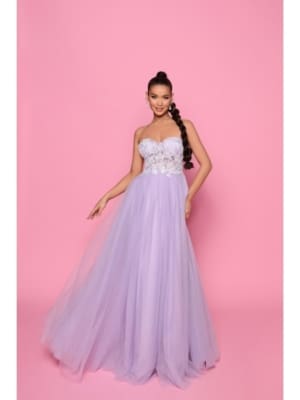 A woman named Suzie stands against a pink background, wearing the SUZIE NP147, a strapless lavender gown featuring a fitted bodice adorned with lace and a flowing tulle skirt. She has long, braided hair and smiles while holding out the skirt of her dress slightly. Angels Formal Wear