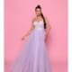 A woman named Suzie stands against a pink background, wearing the SUZIE NP147, a strapless lavender gown featuring a fitted bodice adorned with lace and a flowing tulle skirt. She has long, braided hair and smiles while holding out the skirt of her dress slightly. Angels Formal Wear