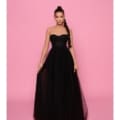 A woman stands against a pink backdrop wearing the SUZIE NP147, a strapless, black, floor-length gown with a fitted bodice and flowing skirt. She has a high, braided ponytail and holds the sides of her dress lightly with both hands, looking directly at the camera. Angels Formal Wear