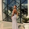 A woman with a graceful updo hairstyle stands on a tiled terrace in the shimmering, backless silver evening gown NAMOI JX4010 from a renowned dress shop. Facing away from the camera, she is positioned near a large window with a crisscross pattern, reflecting the grand architectural beauty of the Sunshine Coast. Angels Formal Wear