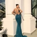 A woman in an elegant, backless NAMOI JX4010 evening gown stands against a white column. The teal dress, perfect for any Dress Shop on the Sunshine Coast, features a long, flowing train. She looks to the side, showcasing her posture amidst modern architectural elements and natural light. Angels Formal Wear