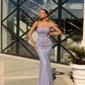 A woman in a shimmery NAMOI JX4010 lavender evening gown stands confidently with one hand on her hip, posing in front of a modern glass building framed by large metal crossbars, reminiscent of those you'd find at a chic Sunshine Coast dress shop, with a decorative white planter and greenery to her right. Angels Formal Wear