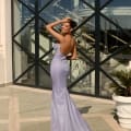 A woman in the dazzling, form-fitting lavender NAMOI JX4010 gown stands on a tiled patio with one hand on her head, perhaps fresh from a visit to the Dress Shop. The dress has a low back and flows into a long train. The background features large glass windows with a diamond pattern and a white exterior railing, capturing the essence of the Sunshine Coast. Angels Formal Wear