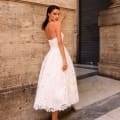 A woman with dark hair, wearing a strapless white lace dress and beige high heels, stands against a stone wall. She looks over her shoulder towards the camera, exuding confidence reminiscent of the PIXIE JX5031. In the background, another stone wall and a closed metal roll-up door complete the scene. Angels Formal Wear