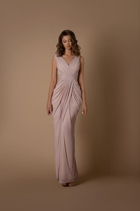 A woman is standing against a plain beige background, wearing the DEMI NBM1001, a long, sleeveless blush-colored gown featuring a draped and pleated design. With wavy hair styled in soft curls and looking down with a serene expression, she exudes the tranquility reminiscent of the Sunshine Coast. Angels Formal Wear