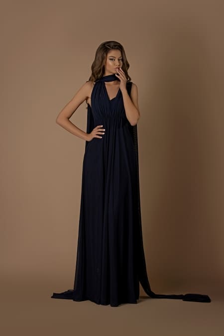 A woman in the NBM1003 long, elegant dark blue gown stands against a neutral background. The dress features a halter neck and drapes gracefully to the floor. She poses with one hand on her hip and the other hand touching her lips, looking to the side, evoking the effortless grace of the Sunshine Coast. Angels Formal Wear