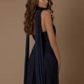 A woman with long, wavy brown hair is posing against a beige background. She is wearing the NBM1003, a dark blue, elegant, backless dress with long ties draped over her shoulders. With a serious expression and a touch of Sunshine Coast allure, she glances to the side. Angels Formal Wear