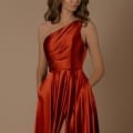 A woman stands against a plain beige background, wearing the shiny, red ALORA NBM1020 one-shoulder dress from Sunshine Coast Dress Shop. The dress features a thigh-high slit and convenient pockets where she rests her hands. Her hair is styled in loose waves, and she confidently gazes at the camera. Angels Formal Wear