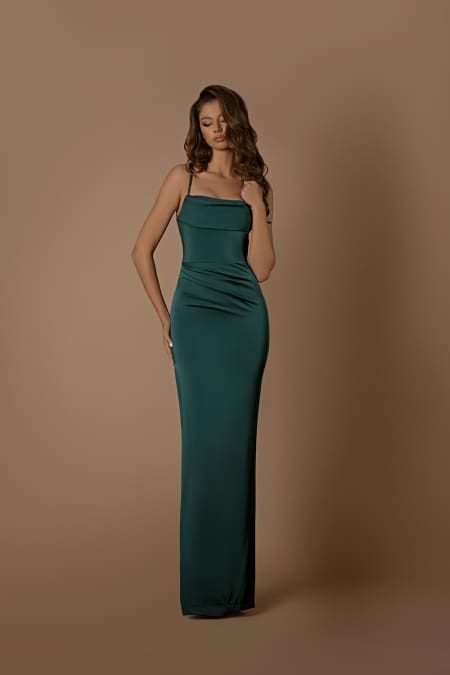 A woman stands against a plain beige background, wearing the dark green, form-fitting, sleeveless KATLYN NBM1021 gown from Sunshine Coast Dress Shop. She has wavy, long brown hair and holds part of her hair with one hand while her other hand rests by her side. She looks down with a serene expression. Angels Formal Wear