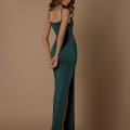 A woman with long, wavy hair poses gracefully, standing sideways and looking down. She dons the sleek and form-fitting KATLYN NBM1021, a sleeveless, floor-length green gown featuring a high slit that reveals her tan high-heeled sandals. Radiating elegance from the Sunshine Coast Dress Shop collection, she stands against a solid light brown background. Angels Formal Wear