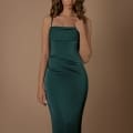 A woman with wavy brown hair is wearing a form-fitting, floor-length emerald green satin dress named KATLYN NBM1021 from Sunshine Coast Dress Shop. She has her hand up near her shoulder strap and poses against a plain beige background. Angels Formal Wear