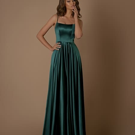 A woman in the ROMANCE NBM1026, a long, flowing dark green satin dress from a chic Sunshine Coast dress store, stands against a plain brown background. The dress features thin straps and a fitted bodice. She is holding her chin with one hand and has loose, wavy hair. Available now with Afterpay options. Angels Formal Wear