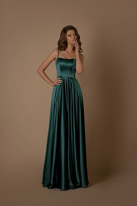 A woman in the ROMANCE NBM1026, a long, flowing dark green satin dress from a chic Sunshine Coast dress store, stands against a plain brown background. The dress features thin straps and a fitted bodice. She is holding her chin with one hand and has loose, wavy hair. Available now with Afterpay options. Angels Formal Wear