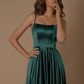 A woman with wavy brown hair stands against a beige background, wearing the ROMANCE NBM1026 long, emerald green satin dress from a Dress Store on the Sunshine Coast. With a contemplative expression, one hand touches her lips while the other rests on her hip. Angels Formal Wear