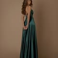 A woman with long, wavy hair stands gracefully against a plain beige background. She is wearing the ROMANCE NBM1026, an elegant, floor-length emerald green satin gown with thin straps and crisscross detailing on the back. Her hands are clasped behind her as she looks over her shoulder, making it perfect for any Dress Store on the Sunshine Coast. Angels Formal Wear