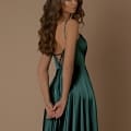 A woman with long, wavy brown hair stands against a plain beige background. She is wearing the ROMANCE NBM1026, a sleeveless deep green satin dress from their favorite Sunshine Coast dress store. The dress features thin straps and a lace-up back detail. She faces away, turning her head slightly to the right while her hands are clasped behind her. Angels Formal Wear