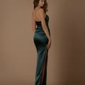 A woman in a strapless, floor-length, dark green satin gown with a high slit on one side poses against a plain brown backdrop. She stands sideways, looking over her shoulder, and has long, wavy hair cascading down her back. Discover similar stunning styles like the MIMI NBM1028 at your favorite Sunshine Coast dress store. Angels Formal Wear