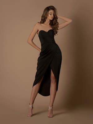 A woman poses against a beige background, wearing the NBM1033, a strapless black mid-length dress with a high slit. She has long, wavy hair and is standing with one hand touching her head and the other on her hip. She is also wearing strappy high heels from Luxury Dress Shop Sunshine Coast. Angels Formal Wear