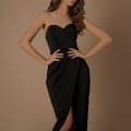 A woman wearing the NBM1033 dress from the Luxury Dress Shop Sunshine Coast, a strapless, black, form-fitting gown with a sweetheart neckline and a thigh-high slit, poses against a plain beige background. She has long, wavy brown hair and stands with her hands resting on her hips. Angels Formal Wear