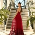 A woman in a flowing, strapless red STELLA NC1003 gown stands at the bottom of an elegant staircase. The dress features intricate embroidery on the skirt. She poses with her back slightly turned, gazing over her shoulder. Lush green plants frame the foreground, with large windows behind her. Angels Formal Wear