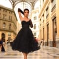 A woman wearing a black strapless dress and matching gloves strikes a stylish pose in an elegant indoor setting featuring high, arched glass ceilings and classical architecture. She stands confidently on the patterned marble floor marked with the product name "NC1004," surrounded by tall columns and expansive windows. Angels Formal Wear