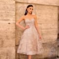 A woman elegantly poses against a textured stone wall, adorned in the NC1004—a strapless, knee-length, beige lace dress featuring intricate floral patterns. She complements her sophisticated look with large hoop earrings, arm-length gloves, and strappy high heels. Angels Formal Wear