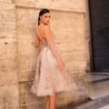 A woman stands against a stone wall with her back to the camera, wearing a light pink, knee-length lace dress with sheer sleeves from NC1004. She complements the dress with nude high heels and large hoop earrings. The background features a closed, dark-colored shutter. Angels Formal Wear