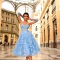 In a grand indoor setting adorned with intricate architecture and towering glass ceilings, a woman stands elegantly. She is wearing the NC1004 light blue strapless lace dress and matching tulle gloves, with her hair styled up as she confidently gazes into the camera. Angels Formal Wear