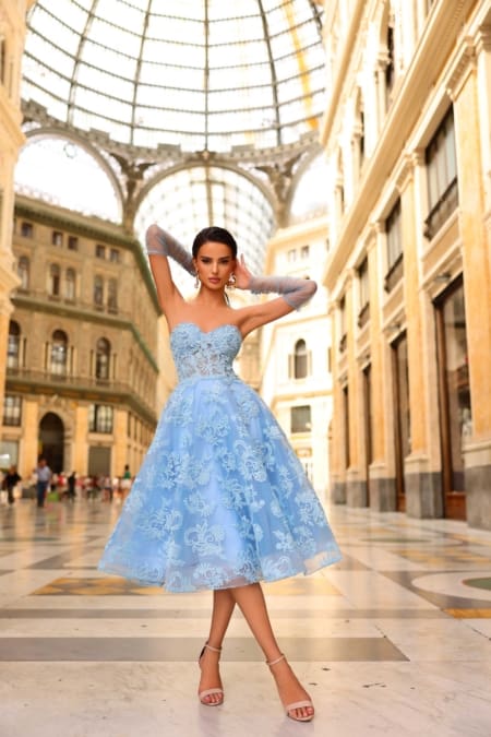 In a grand indoor setting adorned with intricate architecture and towering glass ceilings, a woman stands elegantly. She is wearing the NC1004 light blue strapless lace dress and matching tulle gloves, with her hair styled up as she confidently gazes into the camera. Angels Formal Wear