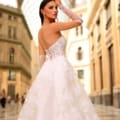 A woman in a white, lace-embroidered strapless dress (Product Name: NC1004) with a corset-style back stands in a grand, ornate architectural setting featuring high glass ceilings and classical details. She faces away, looking over her shoulder with confidence. Angels Formal Wear