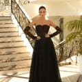 A woman stands confidently at the bottom of an elegant spiral staircase in a dress shop on the Sunshine Coast, wearing the JAZZ NC1005, a floor-length black strapless gown with lace details. Her long gloves match the dress, and her hair is styled in a sleek updo. The setting is luxurious, with ornate iron railings and marble floors. Angels Formal Wear