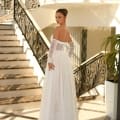 A woman stands on an elegant staircase in a dress shop, turning slightly to look over her shoulder. She is wearing the stunning JAZZ NC1005, a long, white off-the-shoulder gown with intricate lace details and sheer sleeves. The setting features ornate railings and ample natural light, reminiscent of the Sunshine Coast's radiant beauty.
 Angels Formal Wear