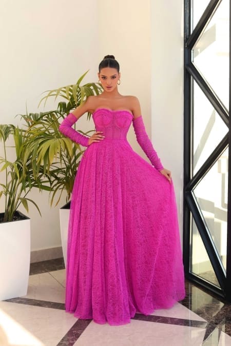 A woman stands indoors at a Sunshine Coast dress shop, wearing the JAZZ NC1005, an off-the-shoulder magenta gown with matching long gloves. The gown features a fitted bodice and a floor-length, flowing skirt. She poses beside large potted plants as natural light streams through the geometric-patterned window. Angels Formal Wear