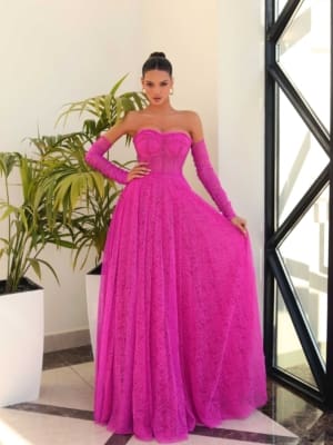 A woman stands indoors at a Sunshine Coast dress shop, wearing the JAZZ NC1005, an off-the-shoulder magenta gown with matching long gloves. The gown features a fitted bodice and a floor-length, flowing skirt. She poses beside large potted plants as natural light streams through the geometric-patterned window. Angels Formal Wear