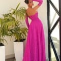 A woman stands next to a window and houseplants, wearing the vibrant pink, off-the-shoulder, full-length JAZZ NC1005 gown from a Sunshine Coast dress shop. She poses with one arm bent, her hand touching her head, showcasing the back of the dress. Angels Formal Wear