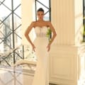 A woman in the NC1007, a strapless, form-fitting white wedding dress with a lace bodice, stands in a sunlit room adorned with tall windows and elegant decor. She has her hands on her hips and is looking directly at the camera. In the background, there is a staircase with an intricate railing, evoking the ambiance of a high-end Sunshine Coast dress shop. Angels Formal Wear