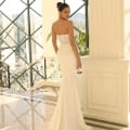 A woman in a strapless, white wedding gown (Product Name: NC1007) with a fitted lace bodice and long train stands inside a bright, spacious dress shop on the Sunshine Coast. Large windows illuminate the shiny marble floor. She looks over her shoulder towards the camera, with elegant railings adorning the staircase behind her. Angels Formal Wear