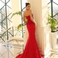 A woman in an elegant NC1007 red strapless gown from a renowned dress shop stands in a bright, sunlit room with large floor-to-ceiling windows and ornate black railings. Her gown has a long train that pools on the floor, and she poses with her back slightly turned, hand resting on a pillar. Angels Formal Wear