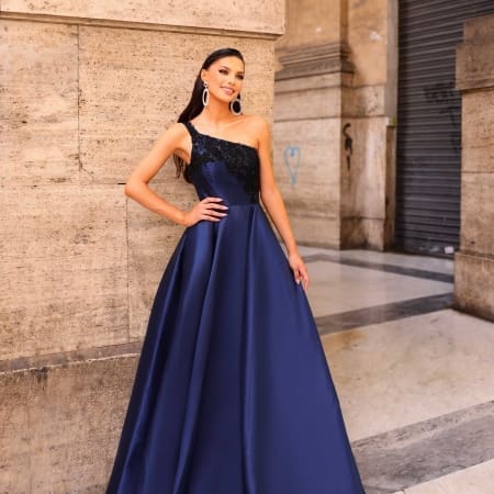 A woman in an elegant, one-shoulder, floor-length navy blue NC1009 gown stands confidently against a stone wall. She has long dark hair and wears large, circular earrings. The setting appears to be an urban, architectural environment with a closed Sunshine Coast Dress Shop door in the background. Angels Formal Wear