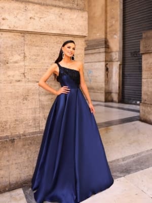 A woman in an elegant, one-shoulder, floor-length navy blue NC1009 gown stands confidently against a stone wall. She has long dark hair and wears large, circular earrings. The setting appears to be an urban, architectural environment with a closed Sunshine Coast Dress Shop door in the background. Angels Formal Wear