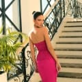 A woman in the NC1010 bright pink, strapless dress with lace detailing stands on an ornate staircase with black wrought-iron railings and white steps. Her hair is pulled back in a sleek bun as she looks over her shoulder. Large windows flood the Sunshine Coast dress shop with light, and a potted plant is visible in the background. Angels Formal Wear