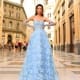 A woman in an intricately embroidered, light blue ball gown, product NC1014 from the Sunshine Coast Dress Shop, stands confidently in an elegant, expansive atrium with a glass ceiling. People are seen walking in the background, adding to the bustling yet sophisticated atmosphere of the setting. Angels Formal Wear