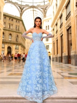 A woman in an intricately embroidered, light blue ball gown, product NC1014 from the Sunshine Coast Dress Shop, stands confidently in an elegant, expansive atrium with a glass ceiling. People are seen walking in the background, adding to the bustling yet sophisticated atmosphere of the setting. Angels Formal Wear