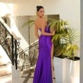 A woman stands elegantly in an opulent setting with a two-story staircase behind her. She is graced in the ELLA NC1015 gown from Dress Shop Sunshine Coast, a form-fitting, strapless purple masterpiece with a long, flowing train. Her hair is styled in a sleek bun as she gazes over her shoulder, adding a touch of allure to her pose. Angels Formal Wear