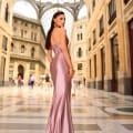 A woman in a floor-length, sleeveless, pink satin gown, product NC1016 from the Sunshine Coast Dress Shop, stands in an elegant, glass-ceilinged gallery adorned with neoclassical architecture. She faces away but glances back slightly with people and architectural details visible in the background. Angels Formal Wear