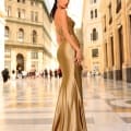 A woman in a long, elegant, shimmering gold dress stands in a grand, ornate arcade with high glass ceilings. She looks over her shoulder, with her hair styled sleek and straight, accessorized with NC1016 drop earrings from Sunshine Coast Dress Shop. The background shows people walking in the distance. Angels Formal Wear