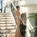 A woman in the ALITZA NC1018, featuring a white lace bodice and a gold satin mermaid skirt, stands on an elegant staircase in a bright, luxurious dress shop on the Sunshine Coast. The staircase features intricate black wrought iron railings, and sunlight illuminates the scene through large windows. Angels Formal Wear